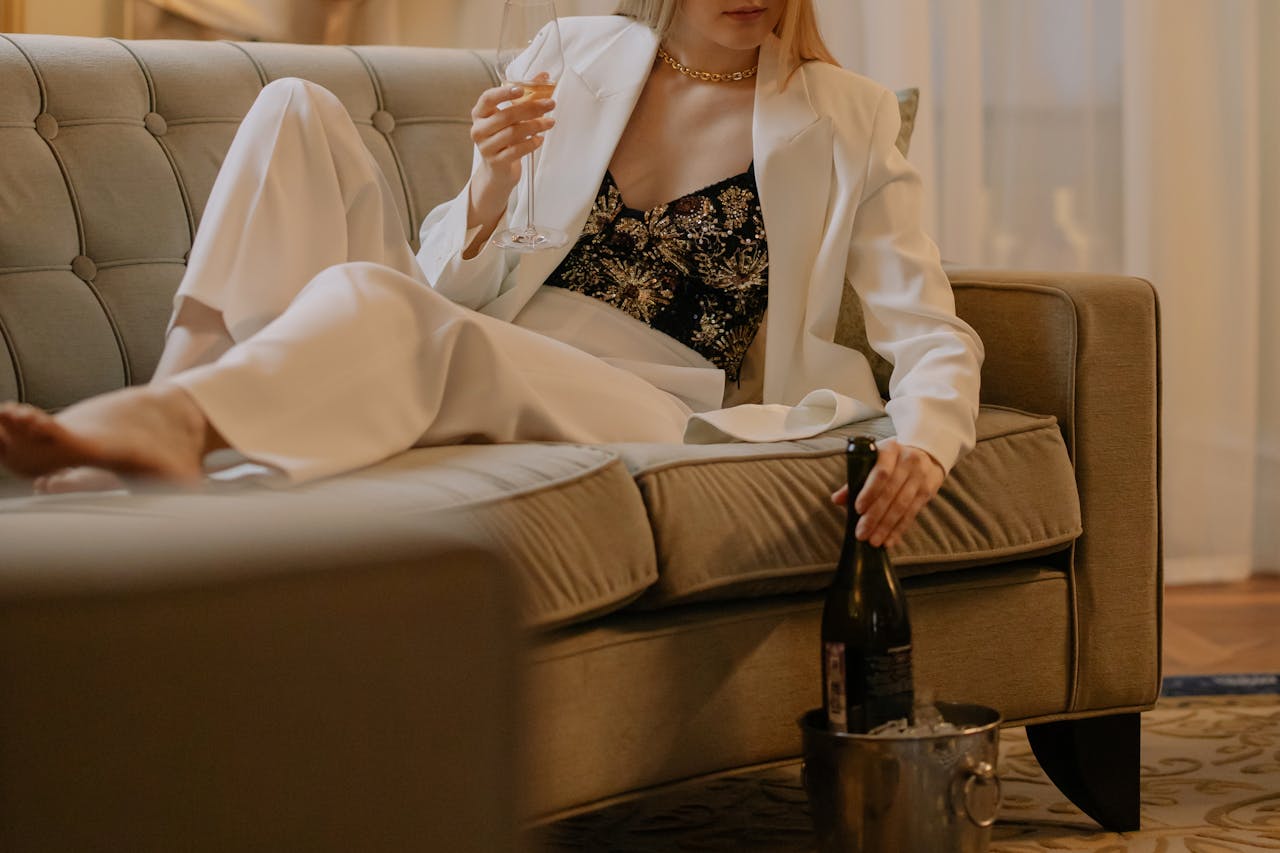Stylish woman in white suit enjoying wine on a sofa in a luxurious hotel room.